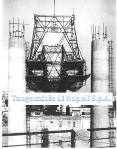 Foto storica: carro-varo a traliccio sospeso tra due pile in costruzione del viadotto, con i palazzi del quartiere sotto